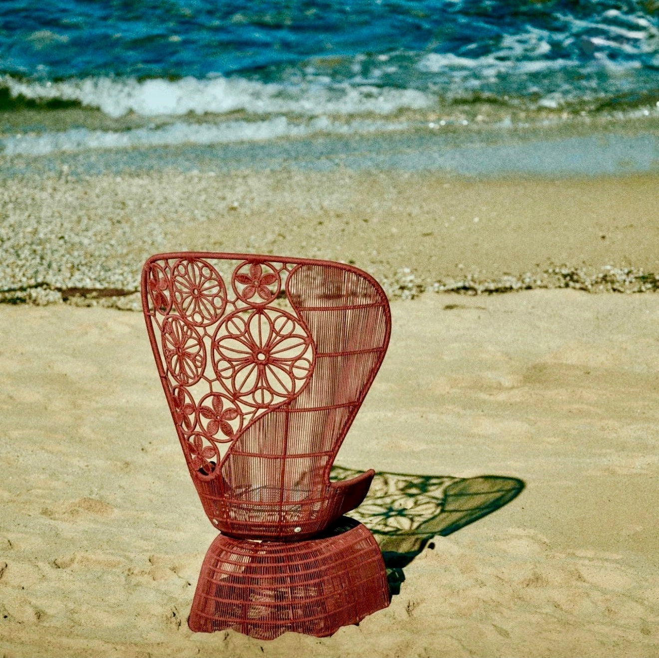 Urban Ashram Genevieve high-back armchair with red woven floral backrest on a sandy beach.